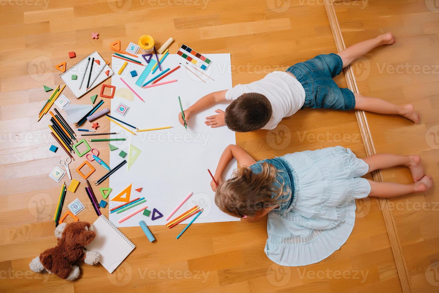 Kids drawing on floor on paper. Preschool boy and girl play on floor with educational toys - blocks, train, railroad, plane. Toys for preschool and kindergarten. Children at home or daycare. Top view. photo