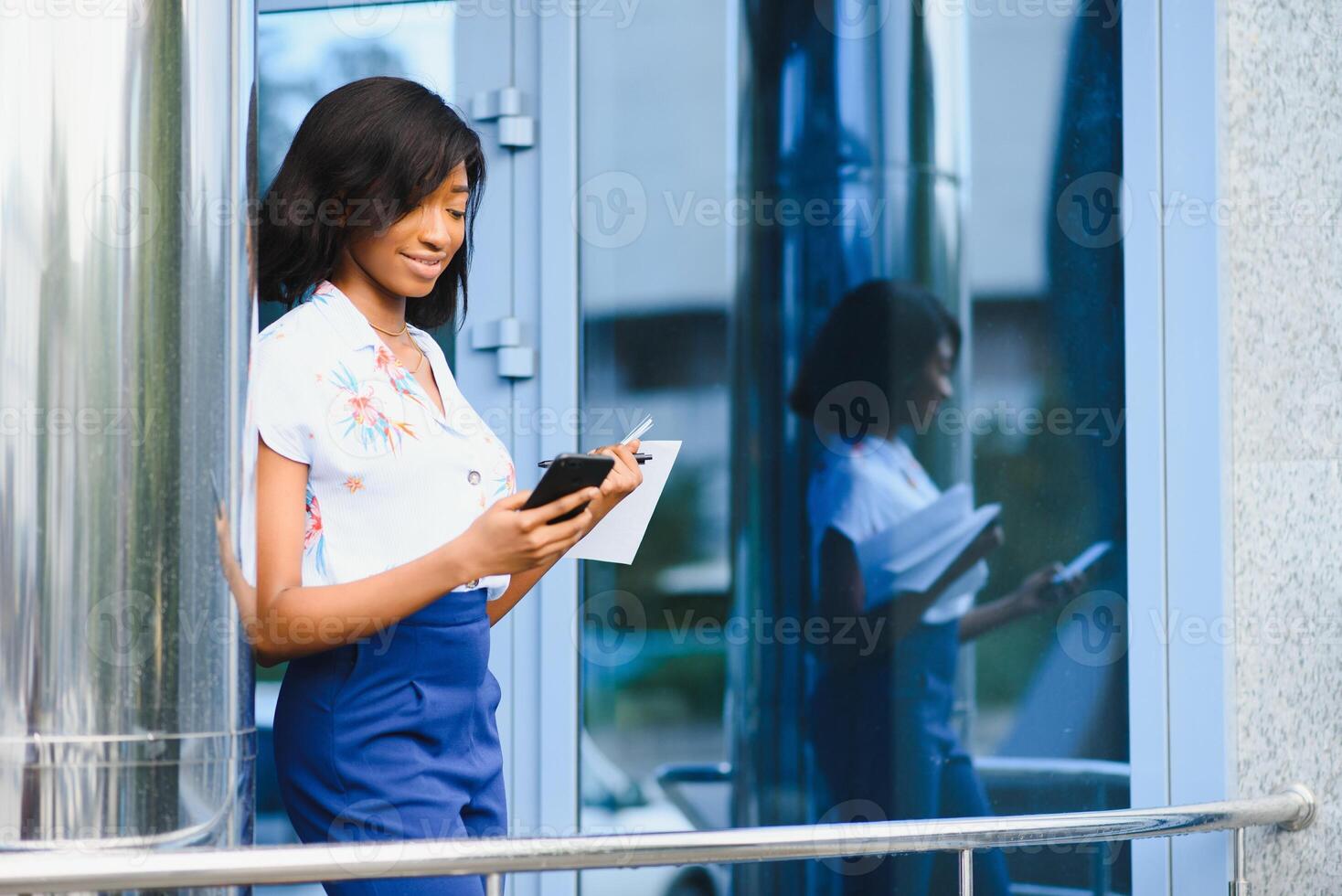 ver desde abajo - linda africano americano niña mecanografía texto en inteligente teléfono en pie en el medio de grande negocio centrar en el ciudad en verano día foto