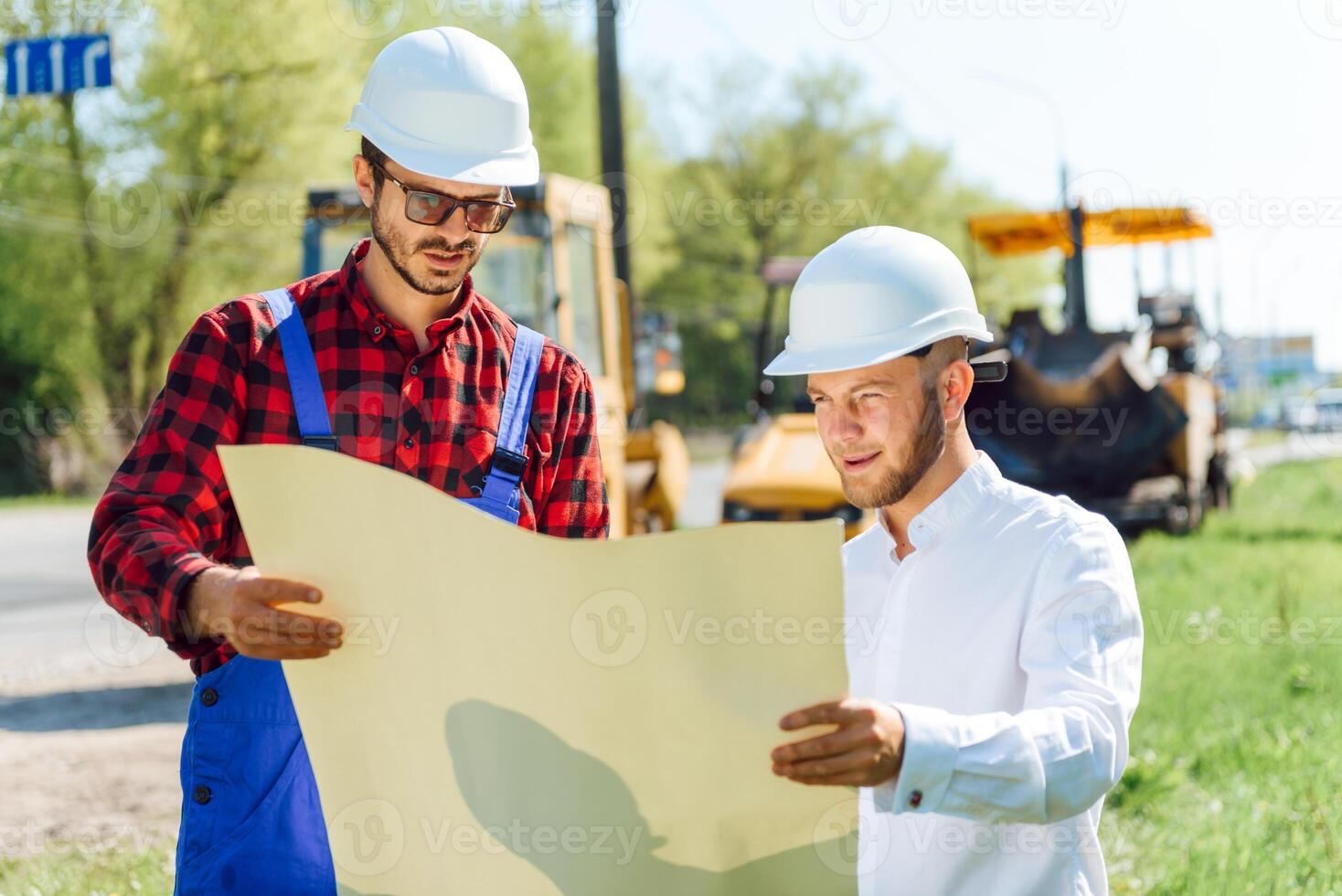 Smiling engineer with helmet standing in front of excavator on road construction site photo