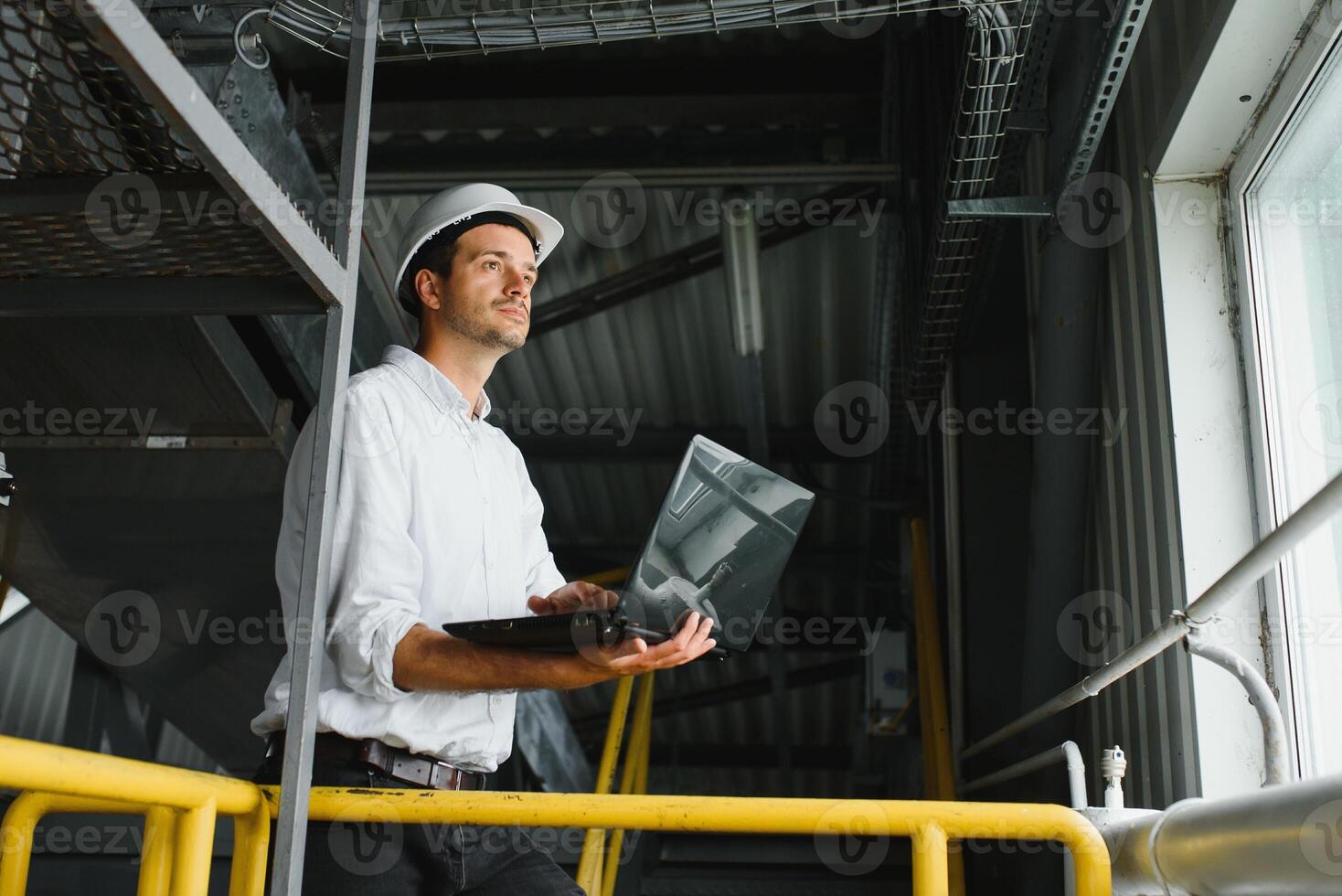 A young handsome employee of a modern factory in a white hard. In the background is a large shop for the production. photo