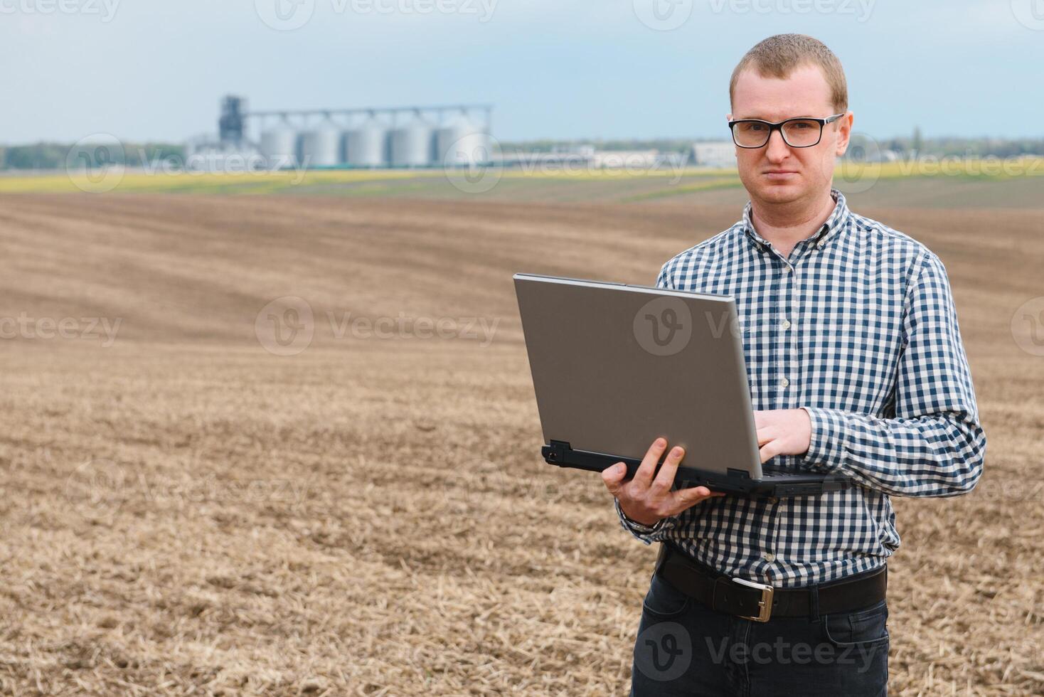 moderno granjero comprobación su campo planta y trabajando en ordenador portátil computadora en contra maíz secadora silos en concepto de industrial y agricultura foto