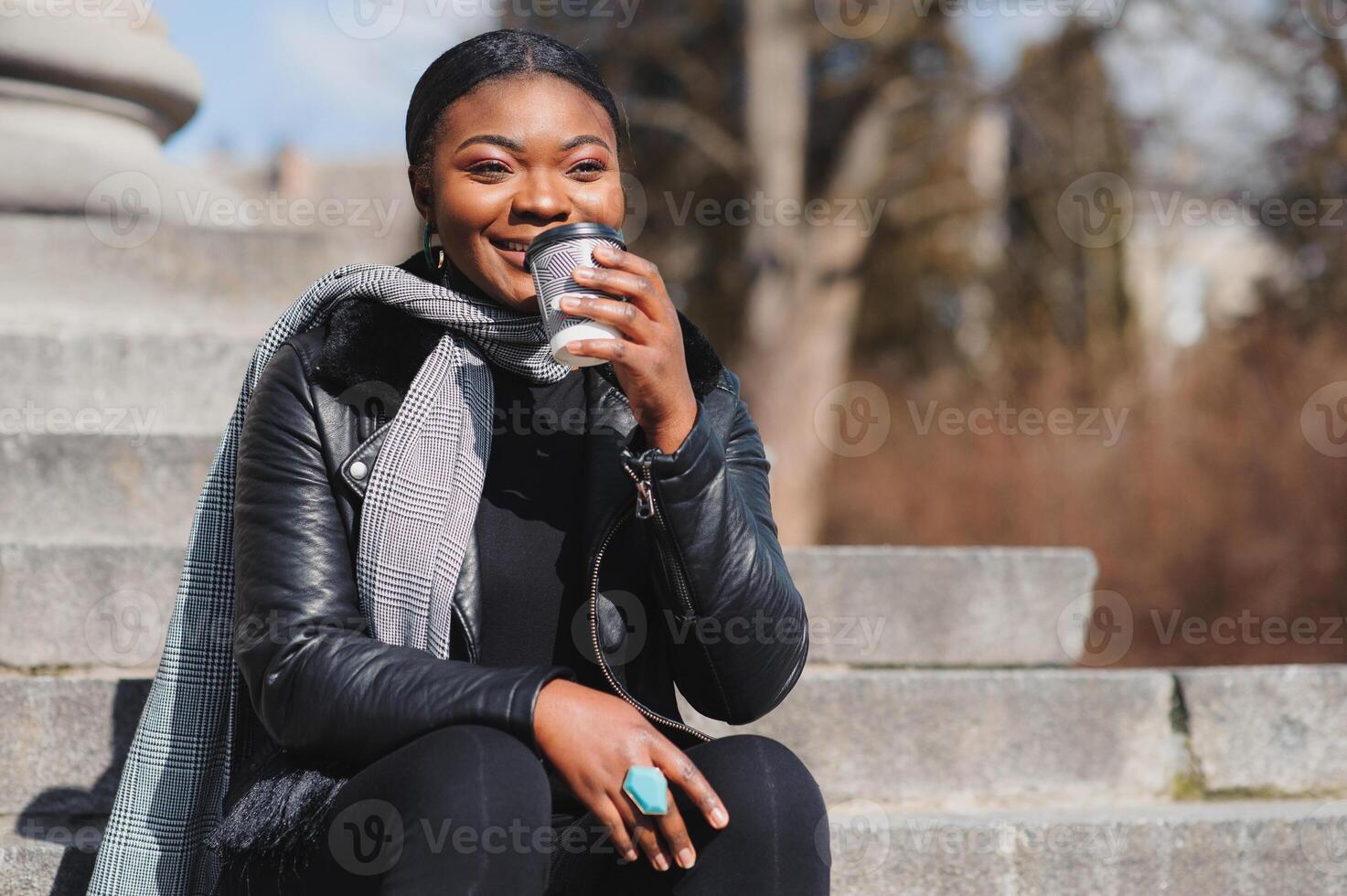 Cheerful dark skinned woman enjoying coffee holding to go cup recreating in city park, happy trendy dressed african american hipster girl sitting outdoors during sunny day drinking beverage photo