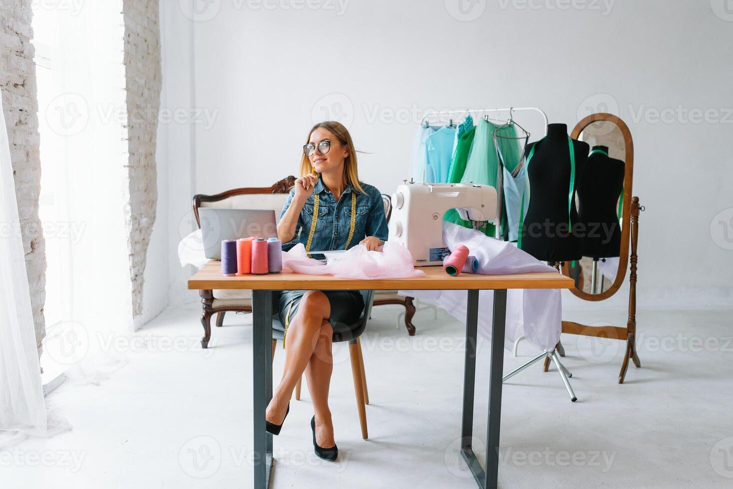 Smiling fashion designer looking at camera at workplace, dressmaker, needlewoman or tailor shop owner sitting at desk with color swatches pantone and embroidery design sketches on the wall, portrait. photo