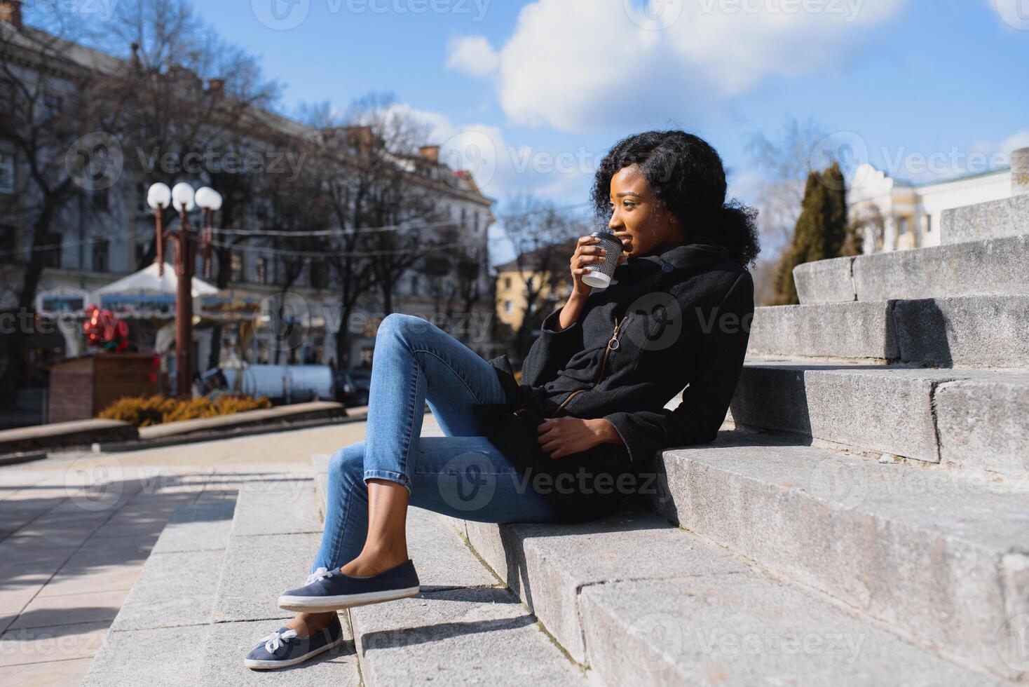 Pensive african woman wearing glasses drinking take away coffee sitting on the street stairs photo