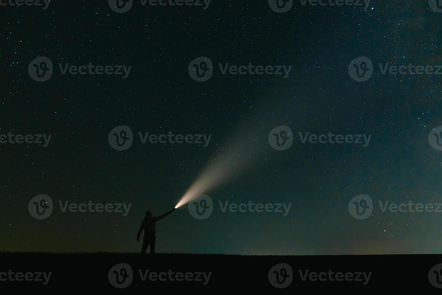 Back view of man with head flashlight standing on green grassy field under beautiful dark blue summer starry sky. Night photography, beauty of nature concept. Wide panorama, copy space background. photo
