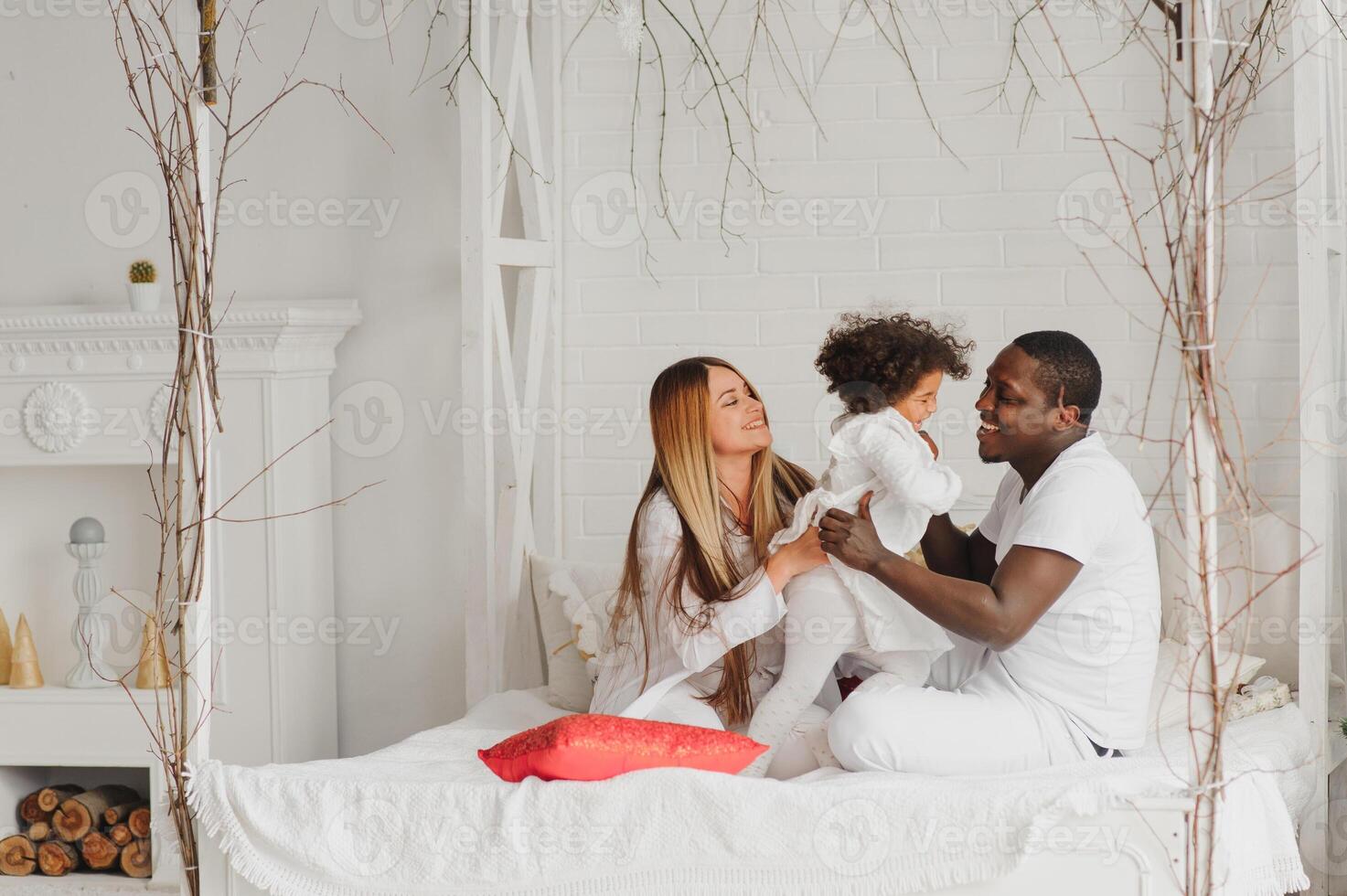 Portrait of happy multiracial young family lying on cozy white bed at home, smiling international mom and dad relaxing with little biracial girl child posing for picture in bedroom photo