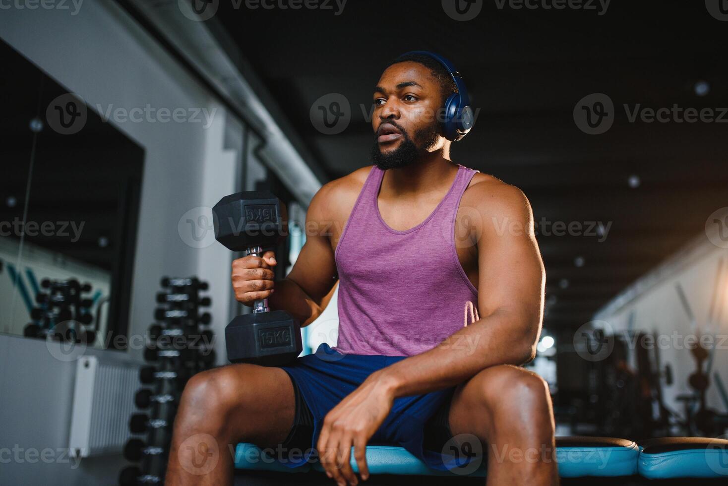 Young African American man sitting and lifting a dumbbell close to the rack at gym. Male weight training person doing a biceps curl in fitness center. photo