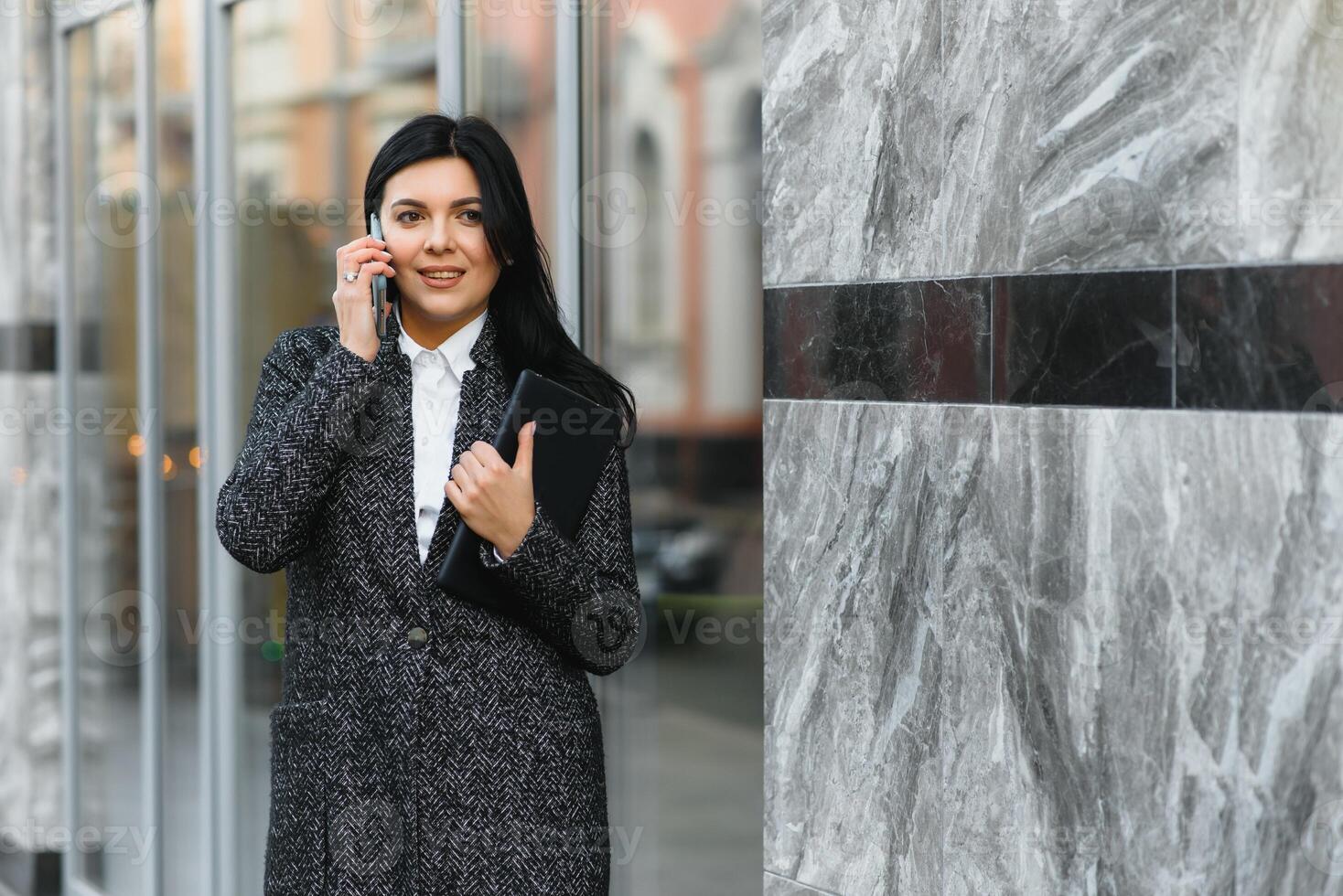 Attractive young woman in the city. Business lady is smiling while talking on a smart phone in front of modern business center photo