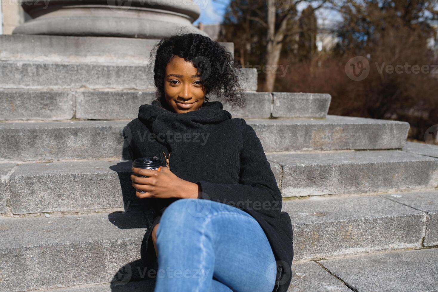 Cheerful dark skinned woman enjoying coffee holding to go cup recreating in city park, happy trendy dressed african american hipster girl sitting outdoors during sunny day drinking beverage photo