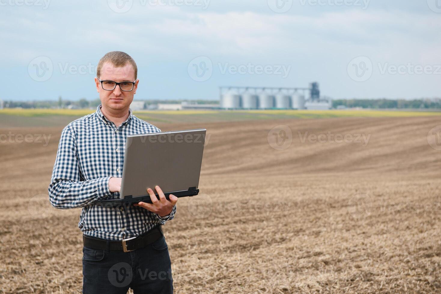 Harvesting concept. farmer in a field with a laptop on a background of a Agricultural Silos for storage and drying of grains, wheat photo