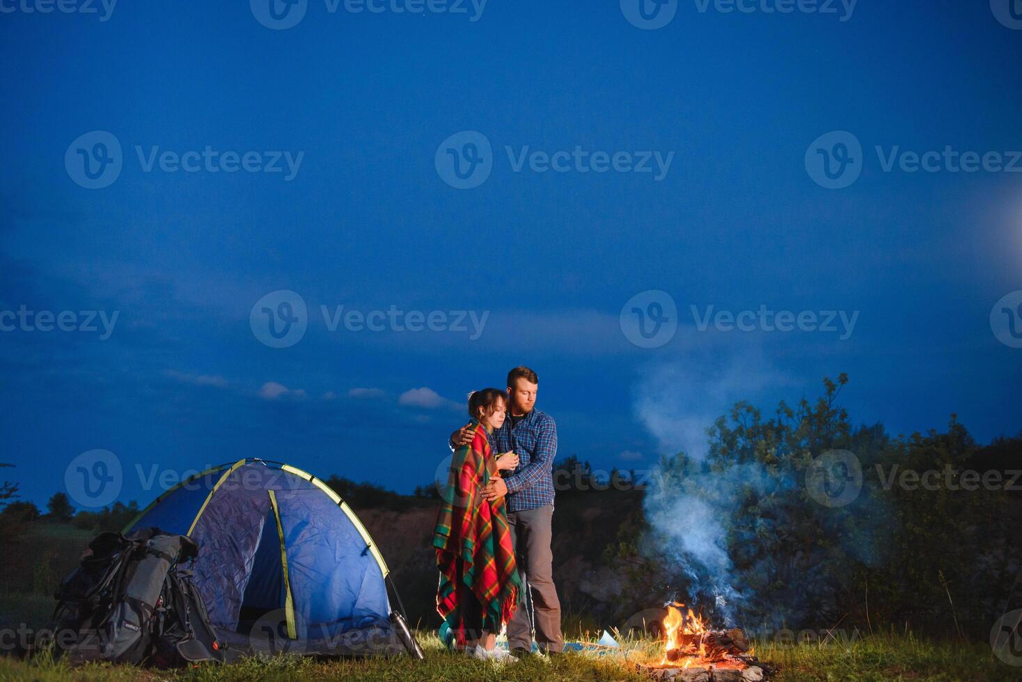 Night camping in the mountains. Happy couple travellers sitting together beside campfire and glowing tourist tent. On background big boulder, forest and night sky. photo