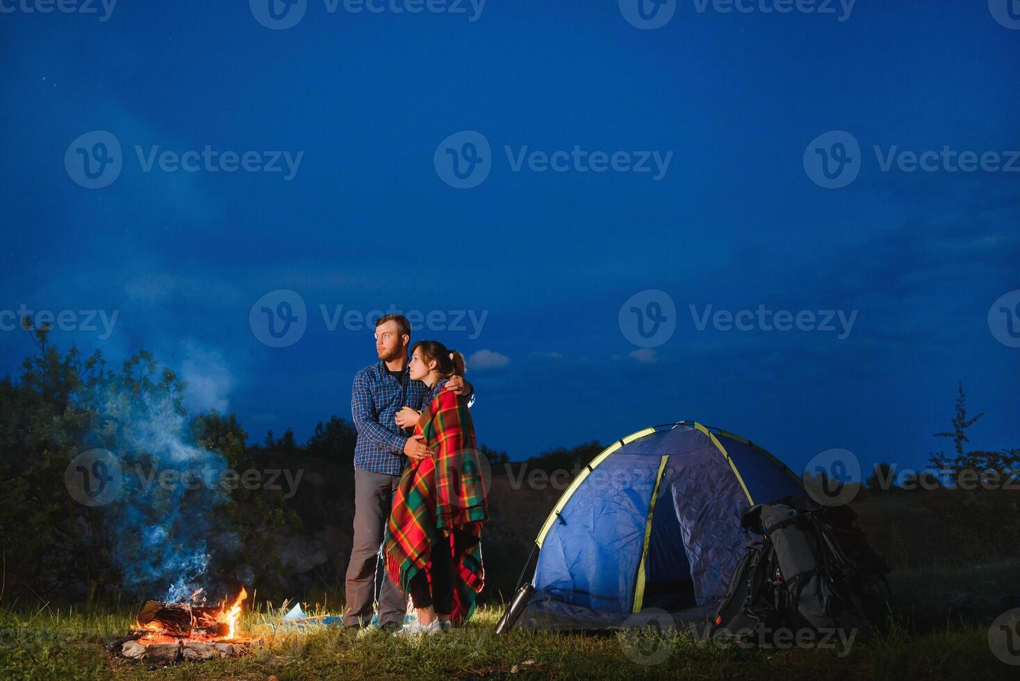 Loving couple hikers enjoying each other, standing by campfire at night under evening sky near trees and tent. Romantic camping near forest in the mountains photo