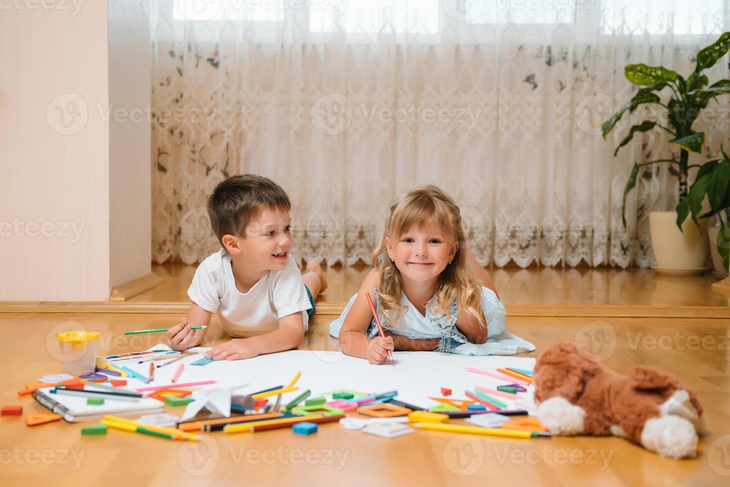 Kids drawing on floor on paper. Preschool boy and girl play on floor with educational toys - blocks, train, railroad, plane. Toys for preschool and kindergarten. Children at home or daycare photo