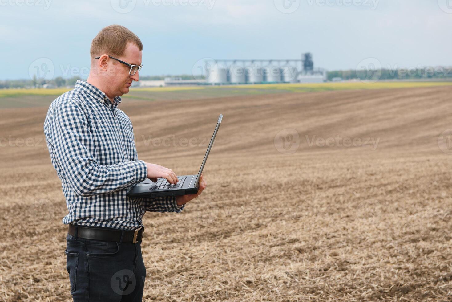 Happy farmer with laptop standing in wheat field in front of grain silo photo