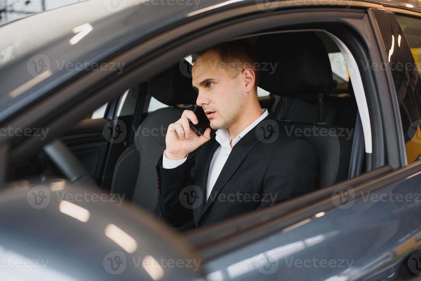 Young businessman sits in luxury car and talks on phone. He looks straight forward. Guy drives car. He holds one hand on steering wheel. It is sunny outside. photo
