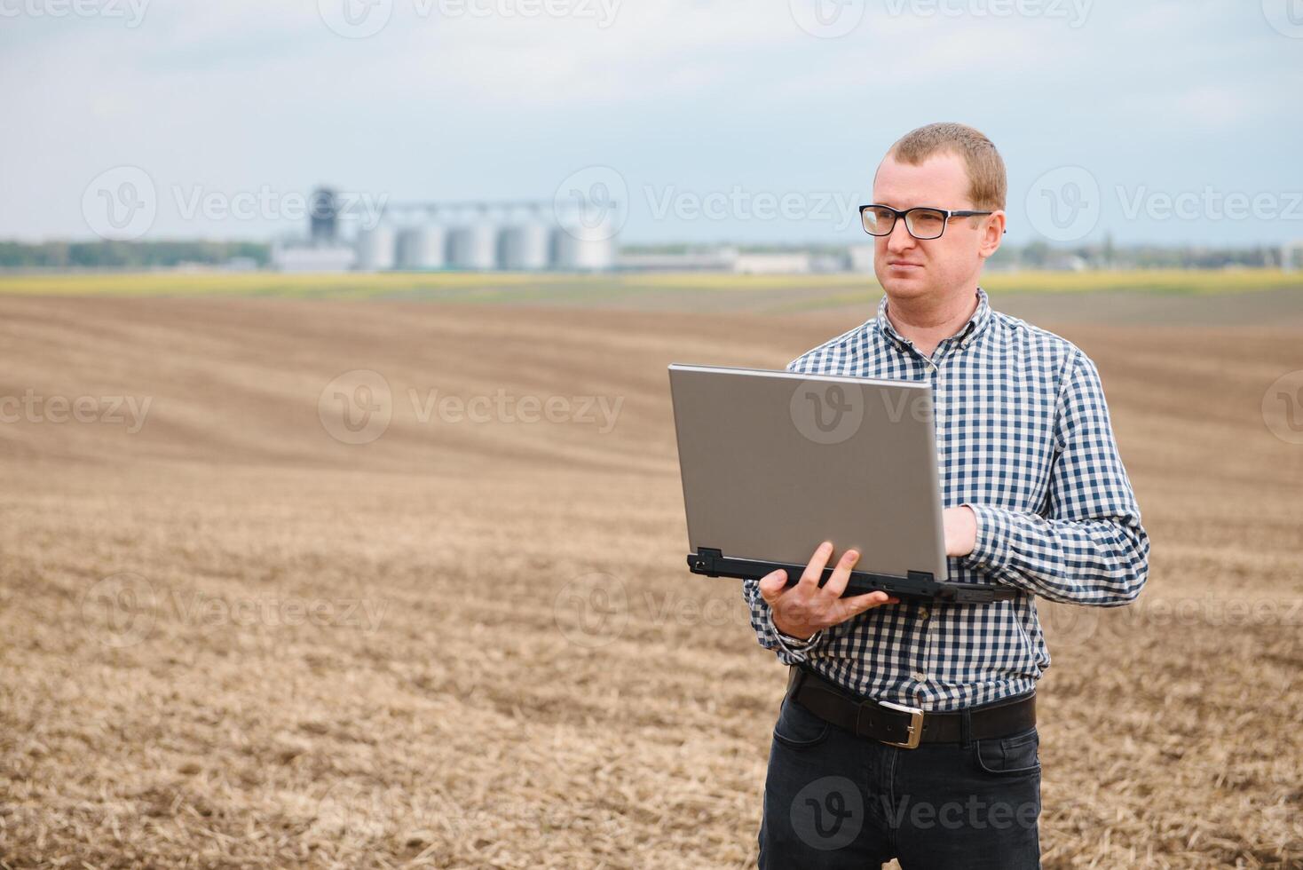 modern farmer checking his field plant and working on laptop computer against corn dryer silos in concept of industrial and agriculture photo