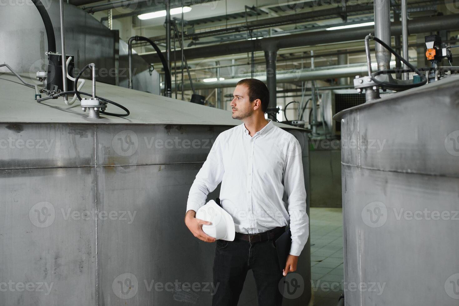 A young handsome employee of a modern factory in a white hard. In the background is a large shop for the production. photo