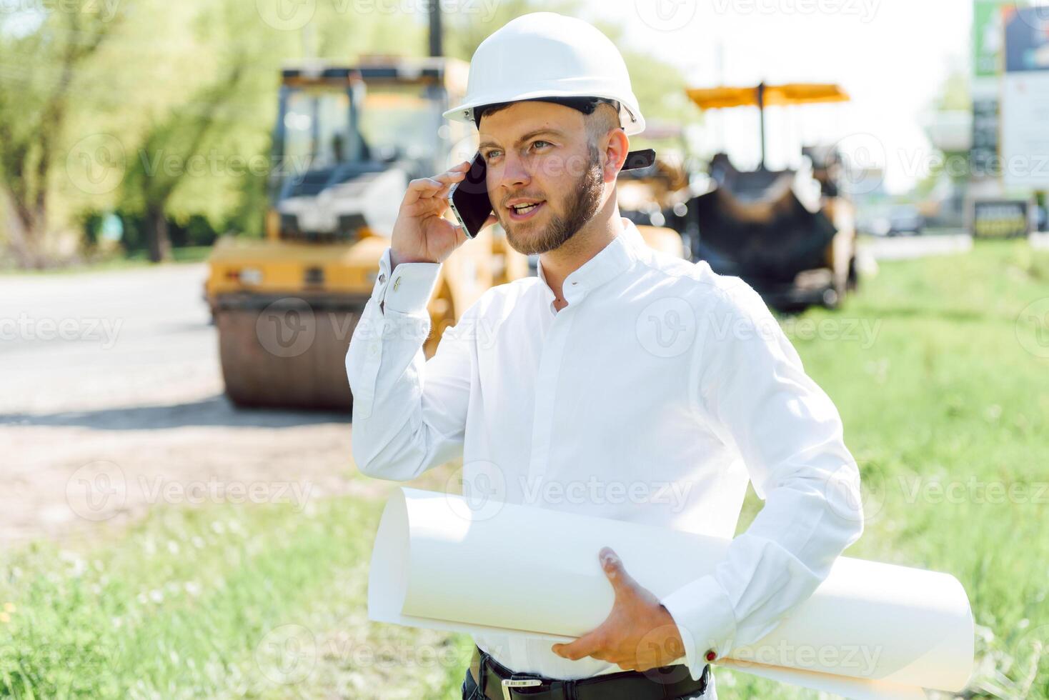 Smiling engineer with helmet standing in front of excavator on road construction site photo