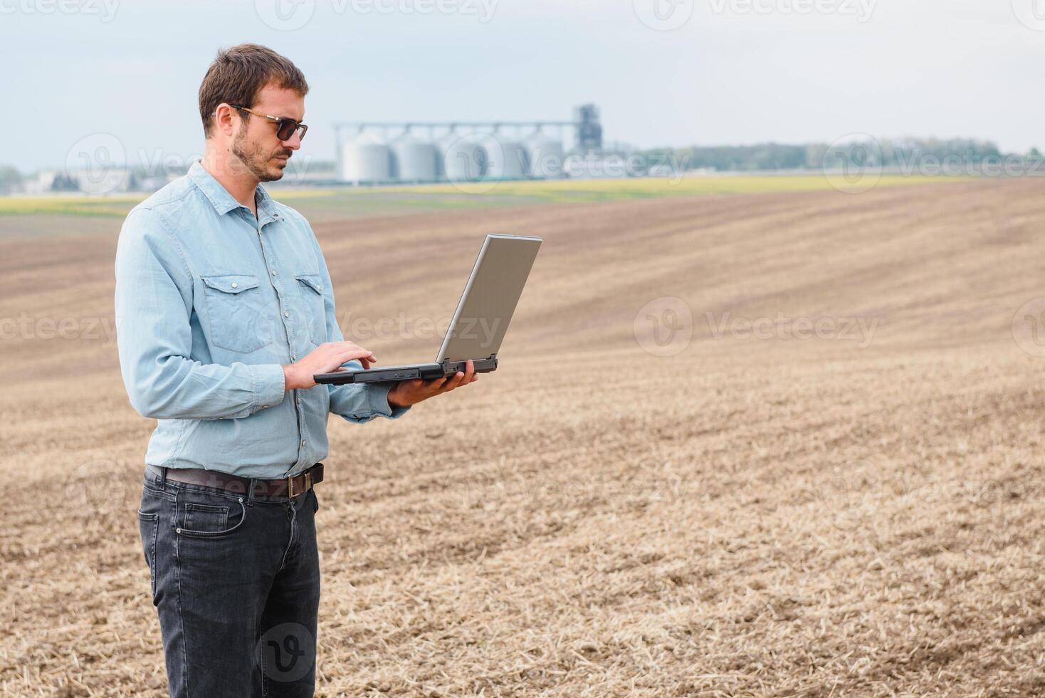 Harvesting concept. farmer in a field with a laptop on a background of a Agricultural Silos for storage and drying of grains, wheat photo