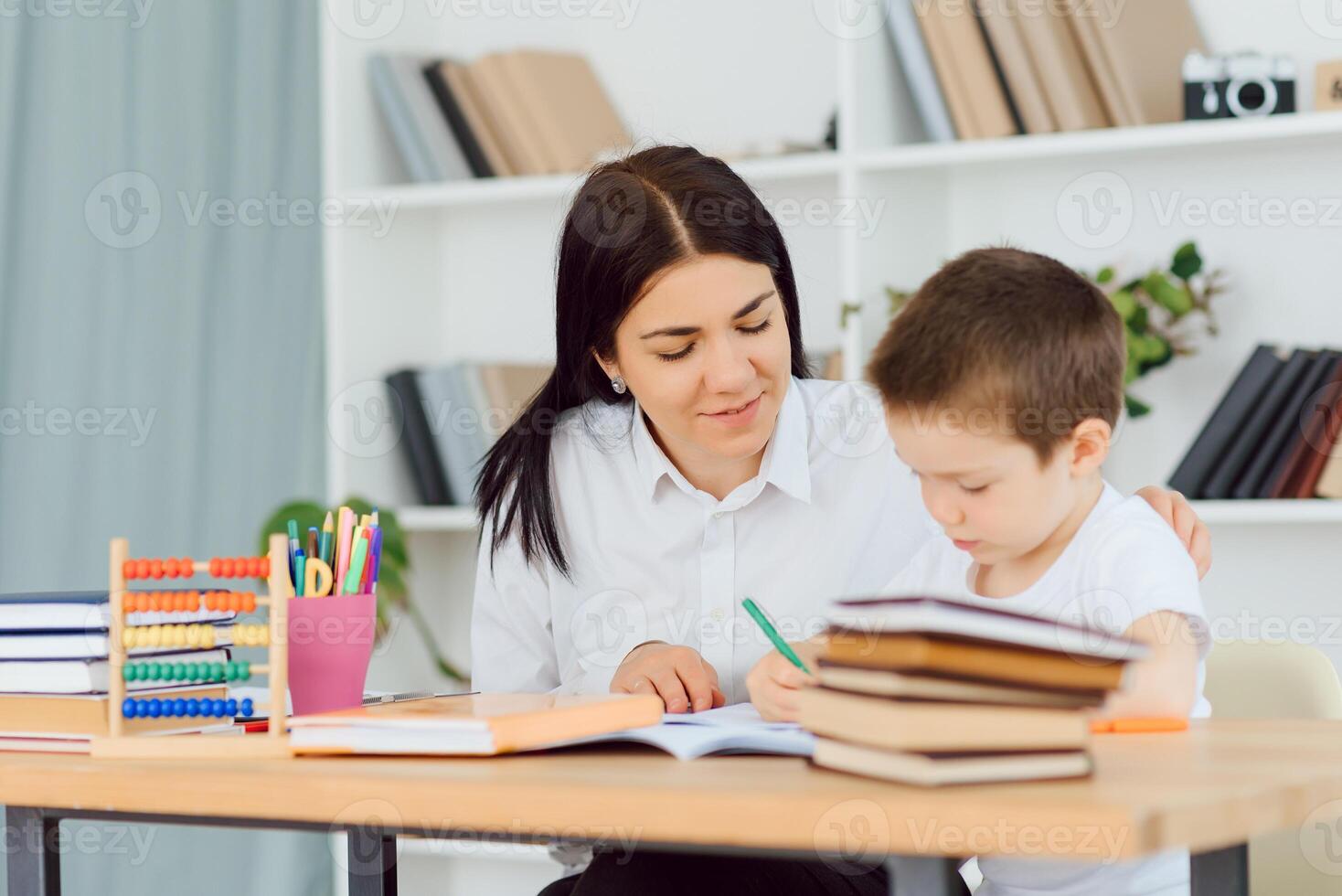 Young female tutor helping little elementary school boy with homework during individual lesson at home photo