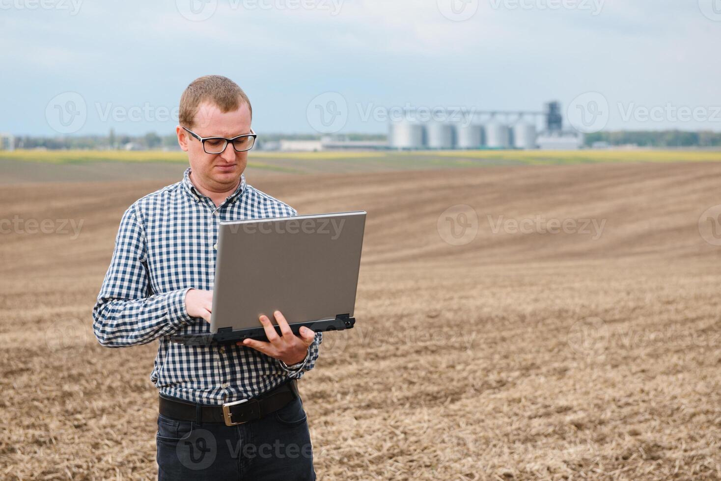 Happy farmer with laptop standing in wheat field in front of grain silo photo