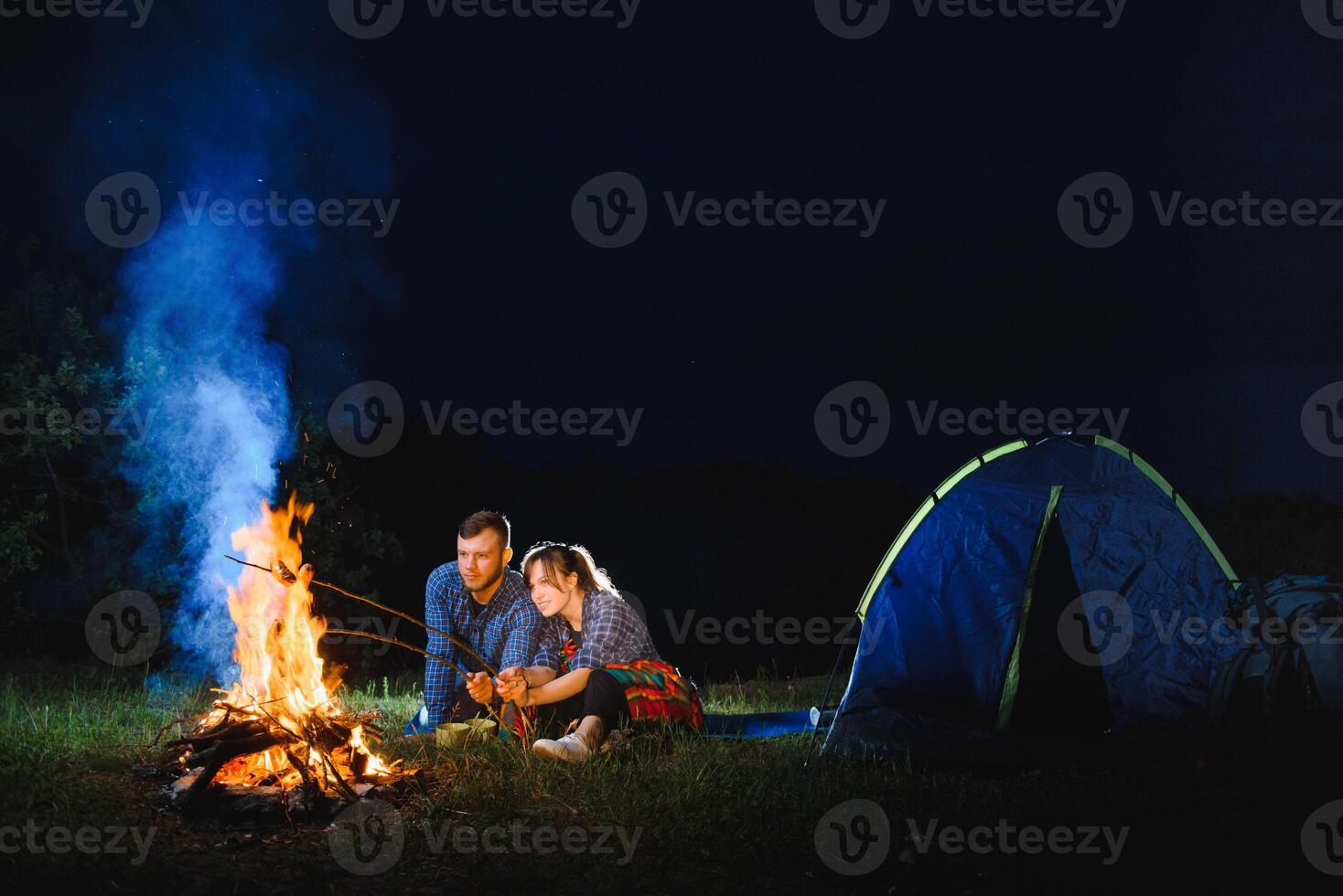 Pareja horneando salchichas en el fuego, y relajante por el hoguera en el bosque en el noche. romántico hoguera concepto. foto