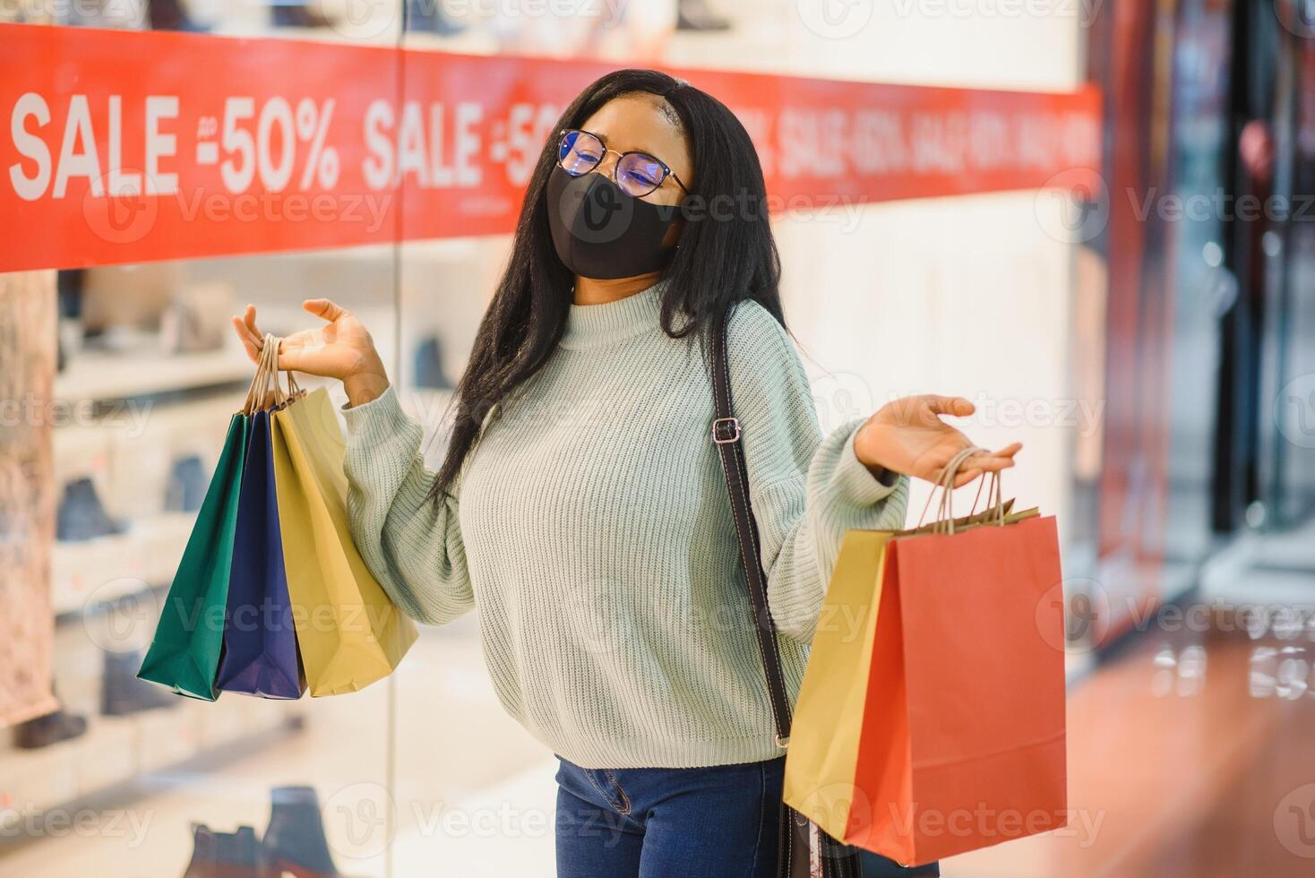 young African American woman at shopping mall in new normal after covid-19 - happy and beautiful black girl in face mask holding shopping bags enjoying at beauty fashion store. photo
