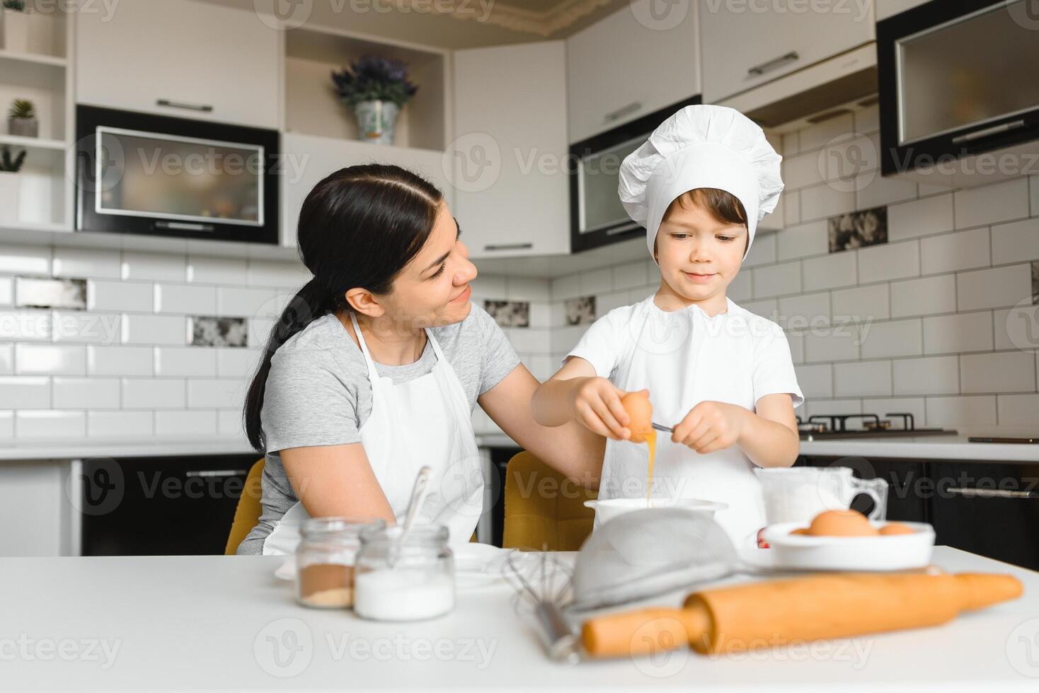 contento madre y su pequeño hijo sacudida crudo huevos en cuenco antes de haciendo masa para hecho en casa Pastelería en el cocina foto