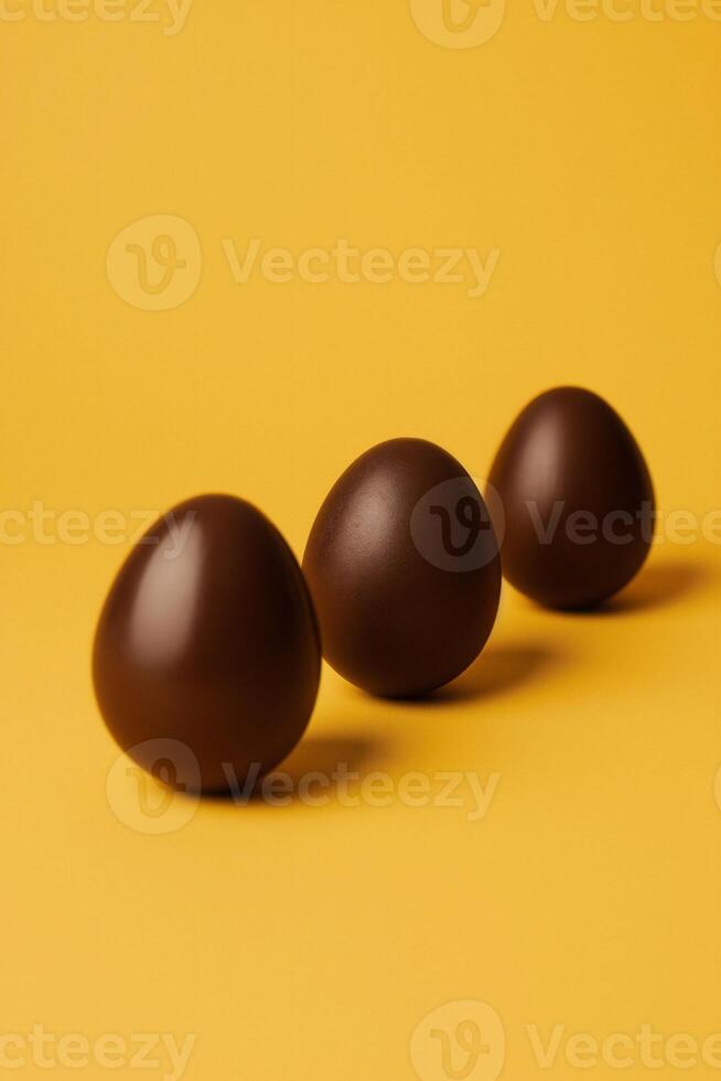 AI generated Three isolated chocolate eggs on a a vibrant yellow background, presenting a bold and minimalistic Easter celebration. Modern marketing campaigns or bold graphic designs photo