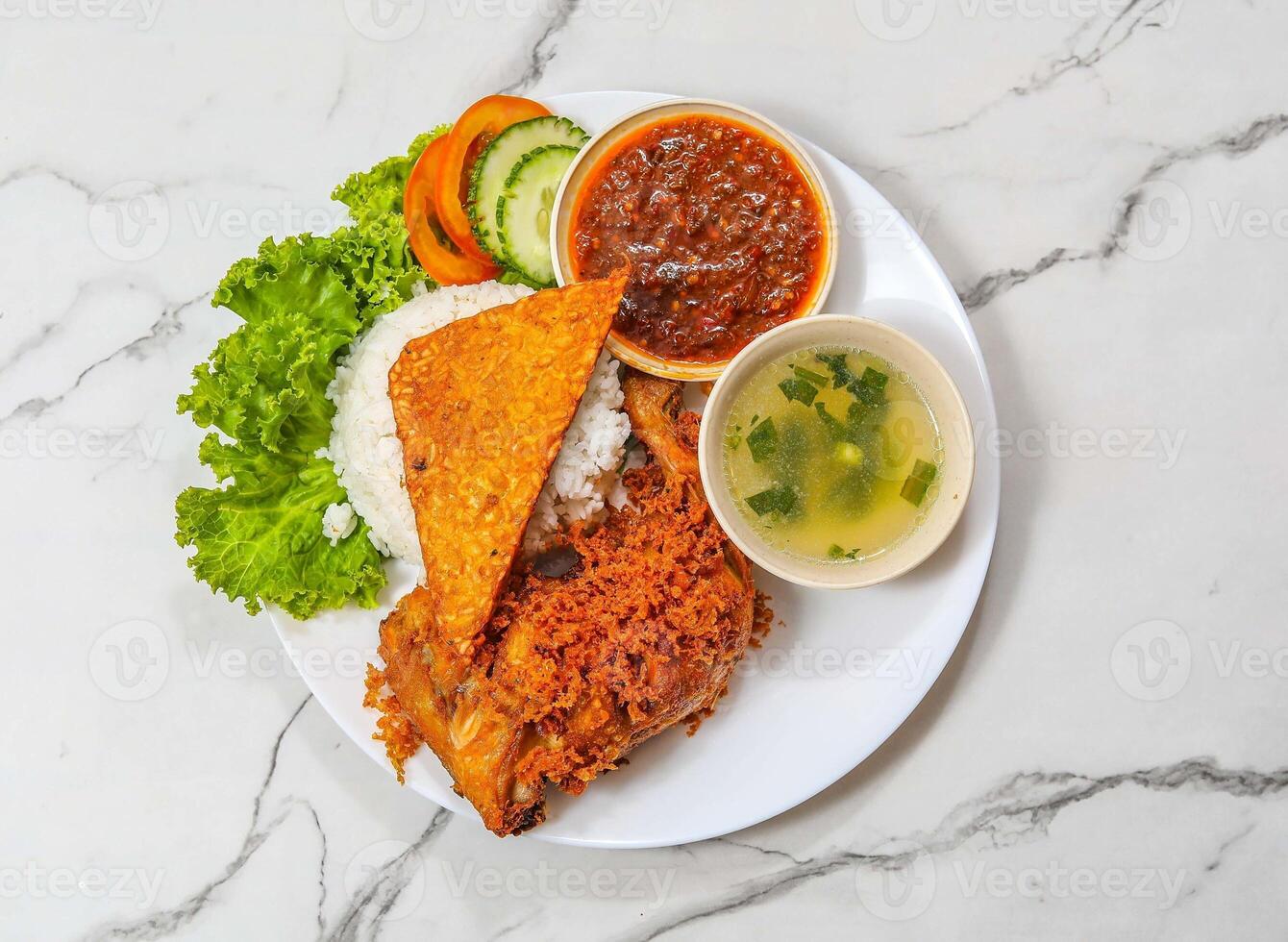 delicious Indonesian food nasi ayam penyet platter with roasted chicken piece, rice, soup, chili sauce and salad isolated on grey marble background top view photo