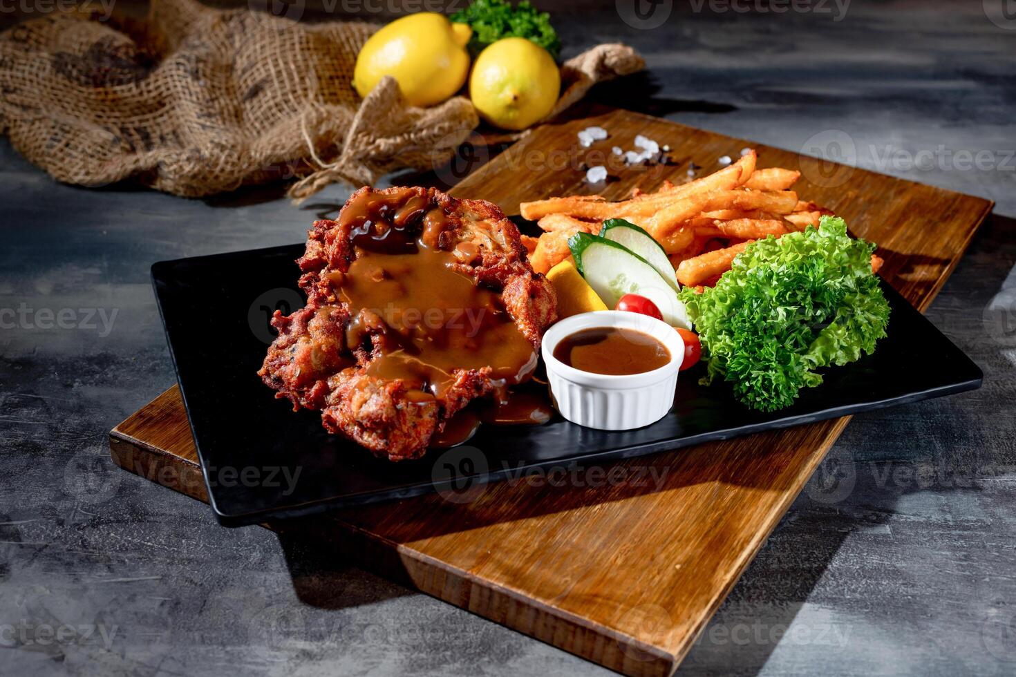 Crispy Boneless Chicken Steak in a black dish with french fries, sauce and lime top view on dark background photo