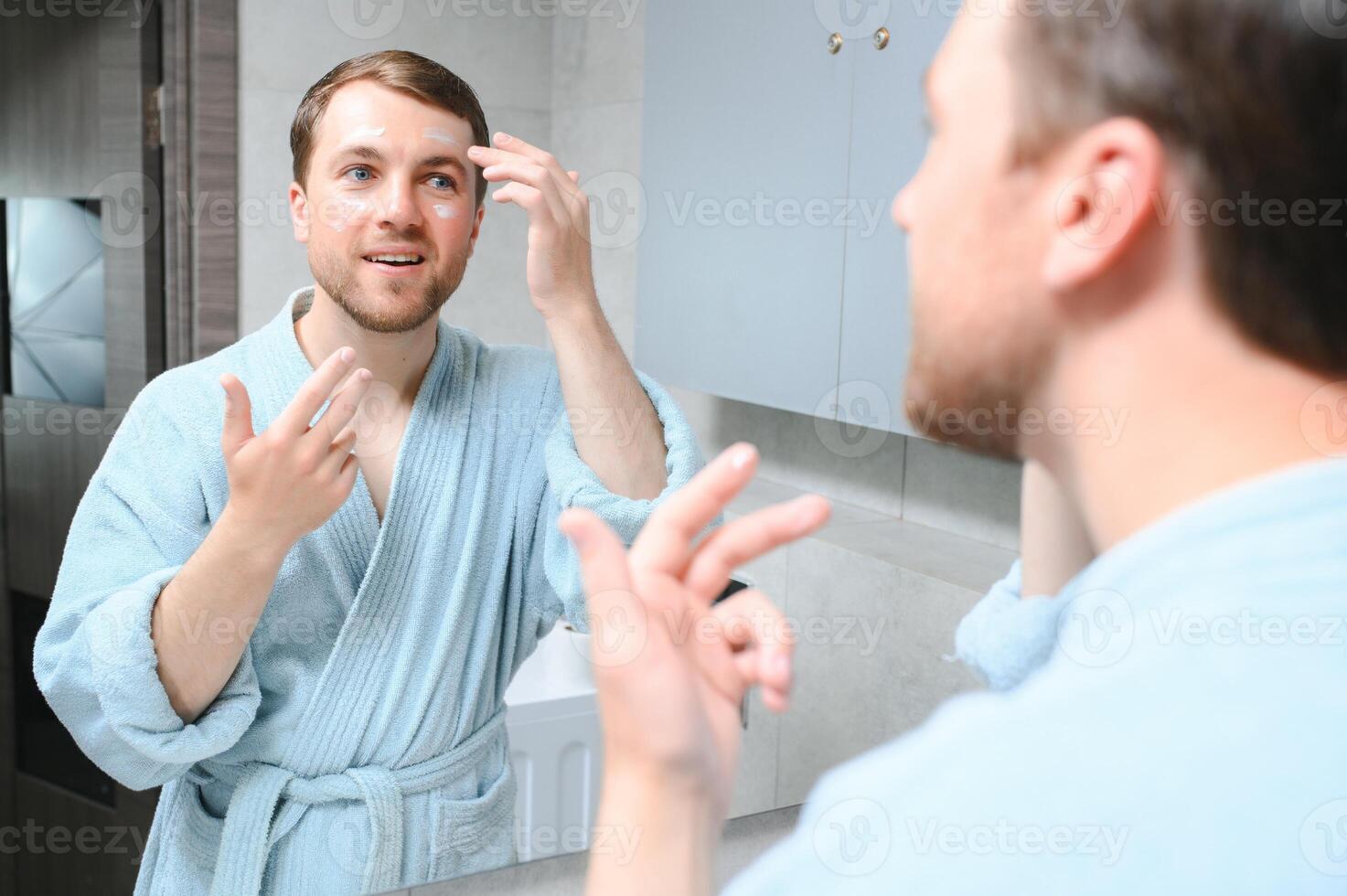 Handsome young man applying cream on his face in bathroom photo