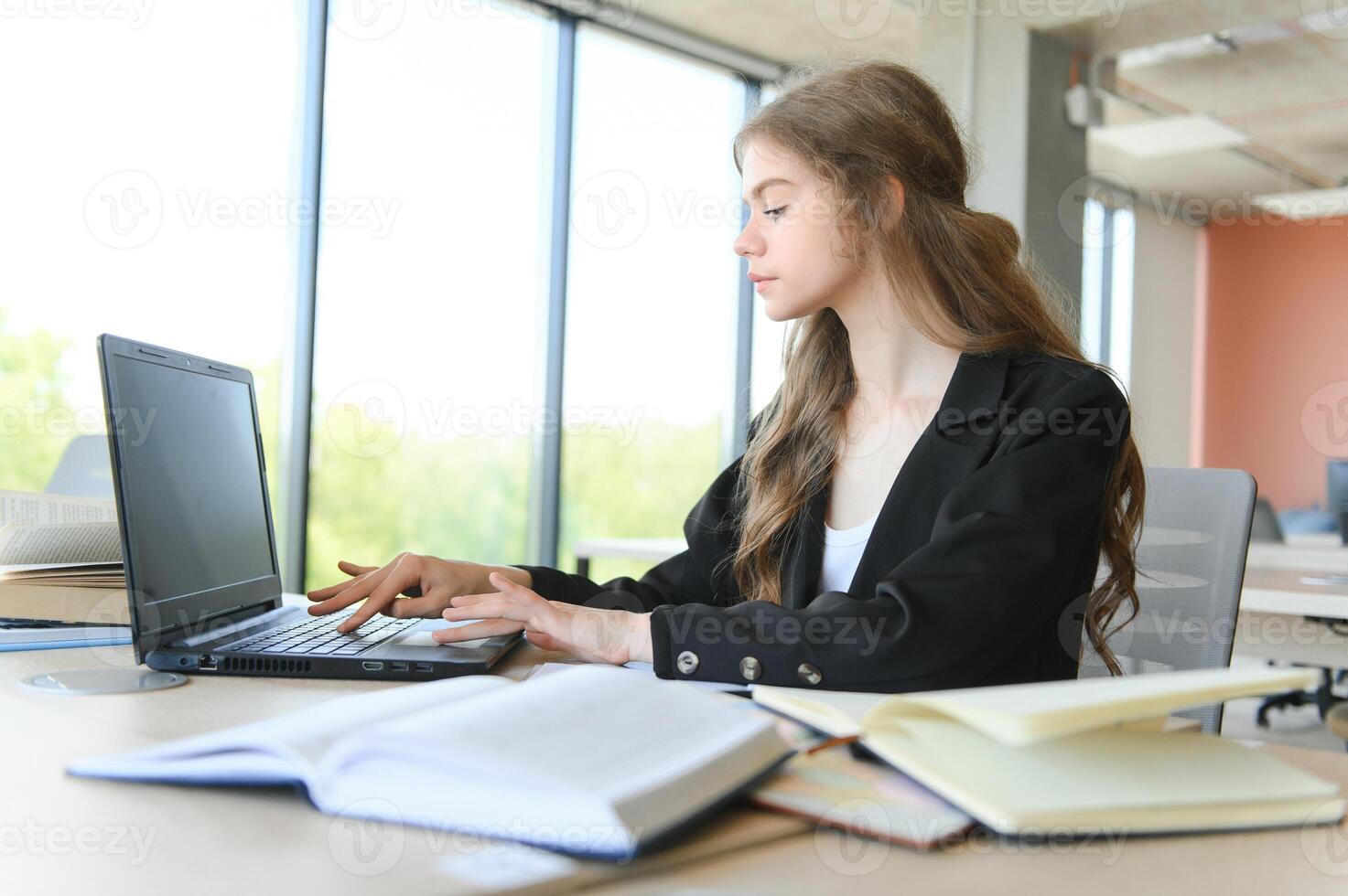 A beautiful caucasian female student is studying in college remotely. She is sitting with a laptop and a notepad and concentrated is watching a video conference lesson photo