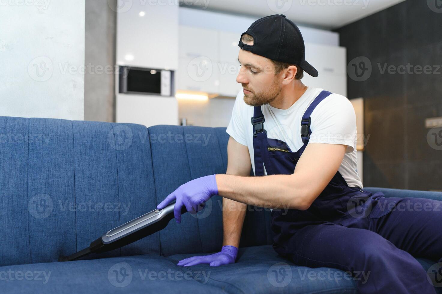 Cleaning service. Man janitor in gloves and uniform vacuum clean sofa with professional equipment. photo