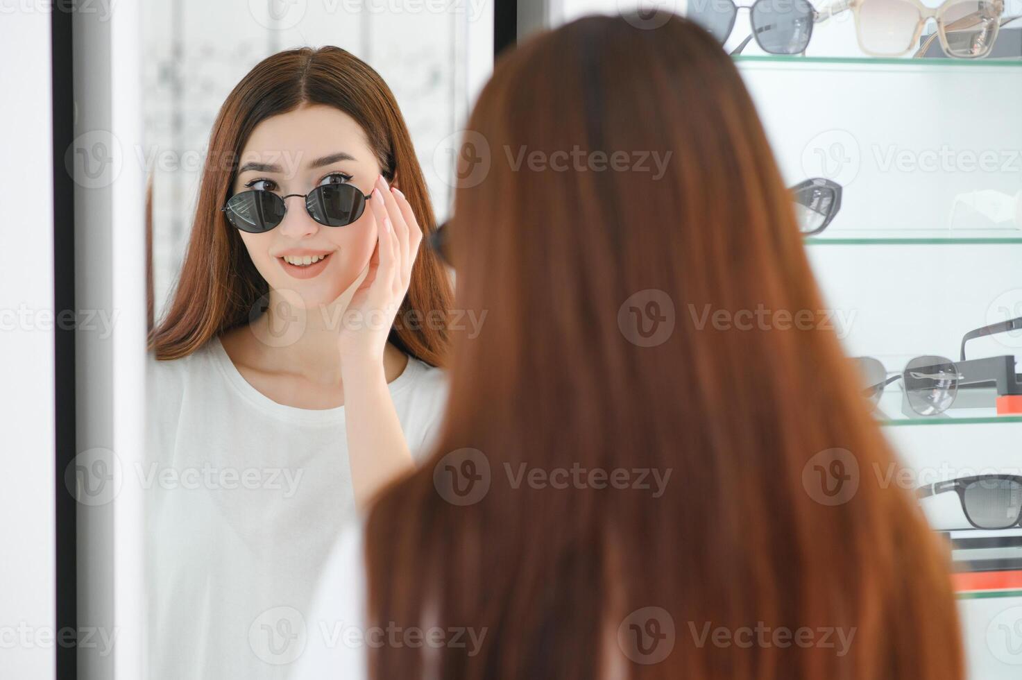 Portrait of a young woman shopping, standing in store and trying sunglasses near a mirror. photo