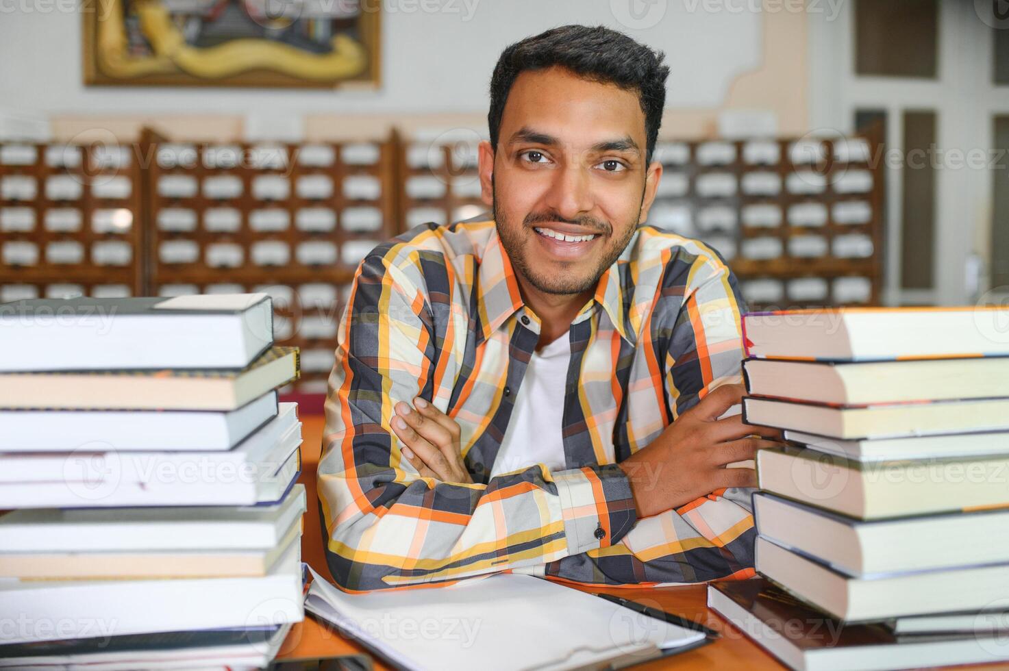 Portrait of cheerful male international Indian student with backpack, learning accessories standing near bookshelves at university library or book store during break between lessons. Education concept photo