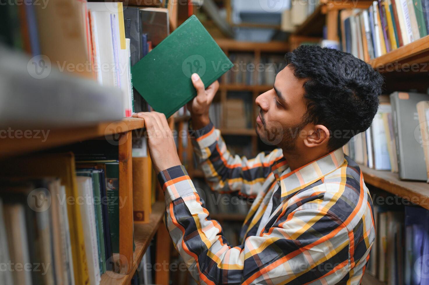 Portrait of cheerful male international Indian student with backpack, learning accessories standing near bookshelves at university library or book store during break between lessons. Education concept photo