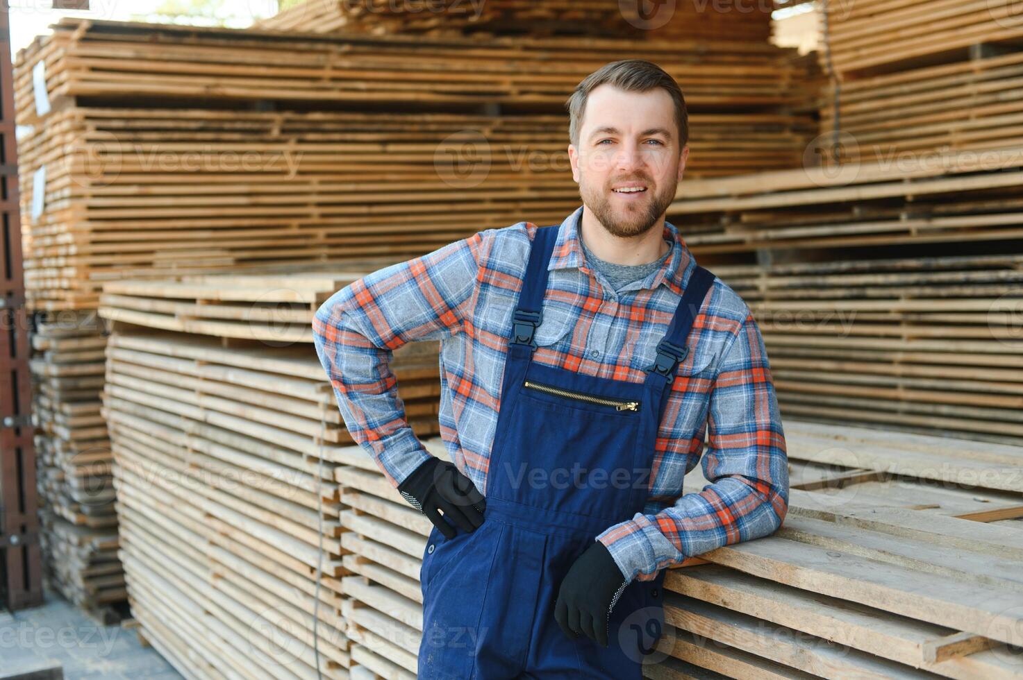Portrait of a handsome worker choosing the best wooden boards. Carpenter standing next to a big stack of wood bars in a warehouse. photo