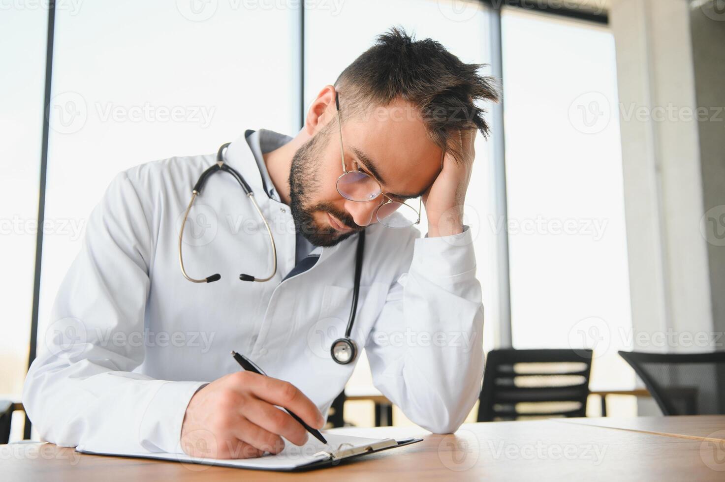 hermoso médico hombre vistiendo médico uniforme sentado en su lugar de trabajo cansado participación su cabeza sensación fatiga y dolor de cabeza. estrés y frustración concepto foto