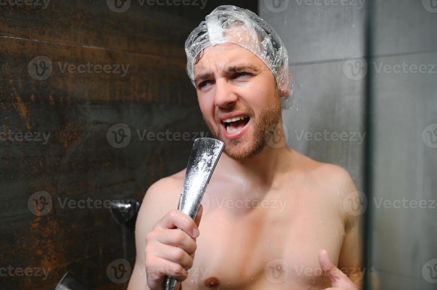 Attractive young cheerful man singing while washing in the shower, wearing shower cap and holding shower head photo