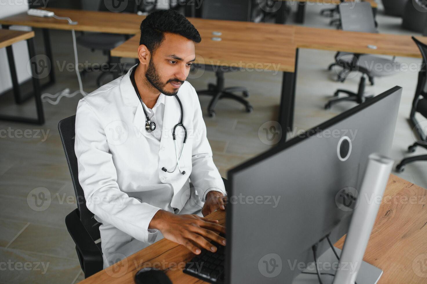 People, occupation and medicine concept. Smiling male Indian Arab doctor in white coat, sitting in medical office at desk with laptop photo