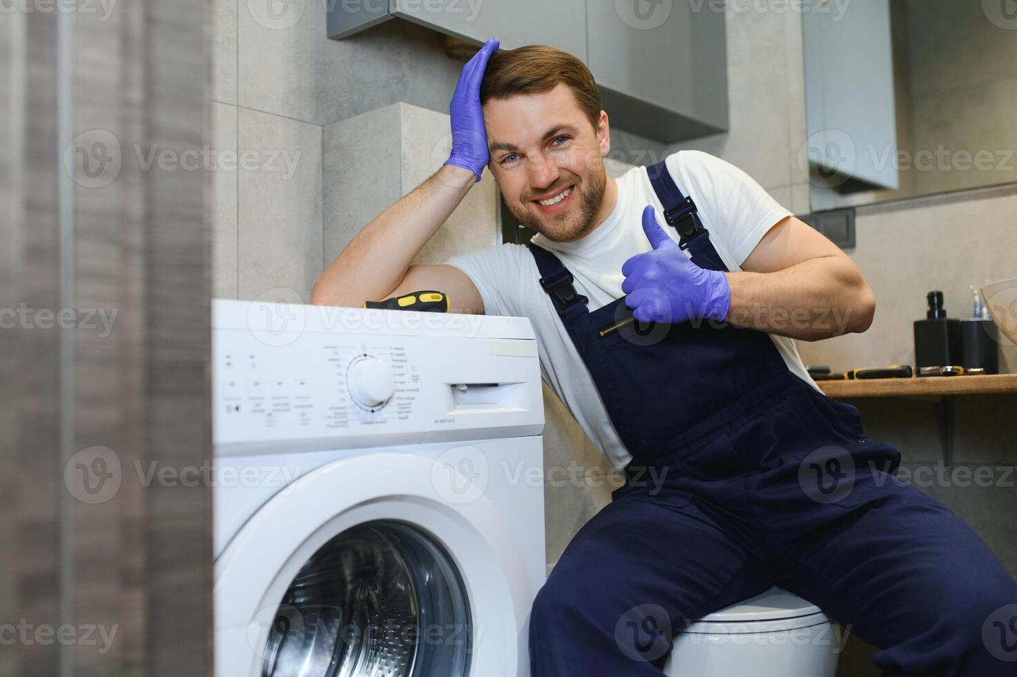 The young handsome repairman in worker suit with the professional tools box is fixing the washing machine in the bathroom photo