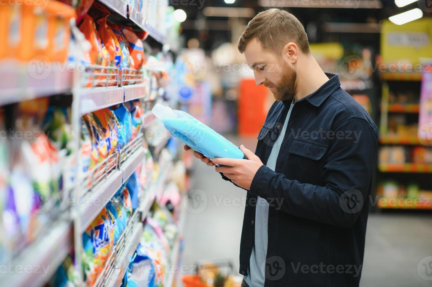 Young man in the supermarket in the household chemicals department. Large selection of products. A brunette in a glasses and a beard in a beige coat. photo