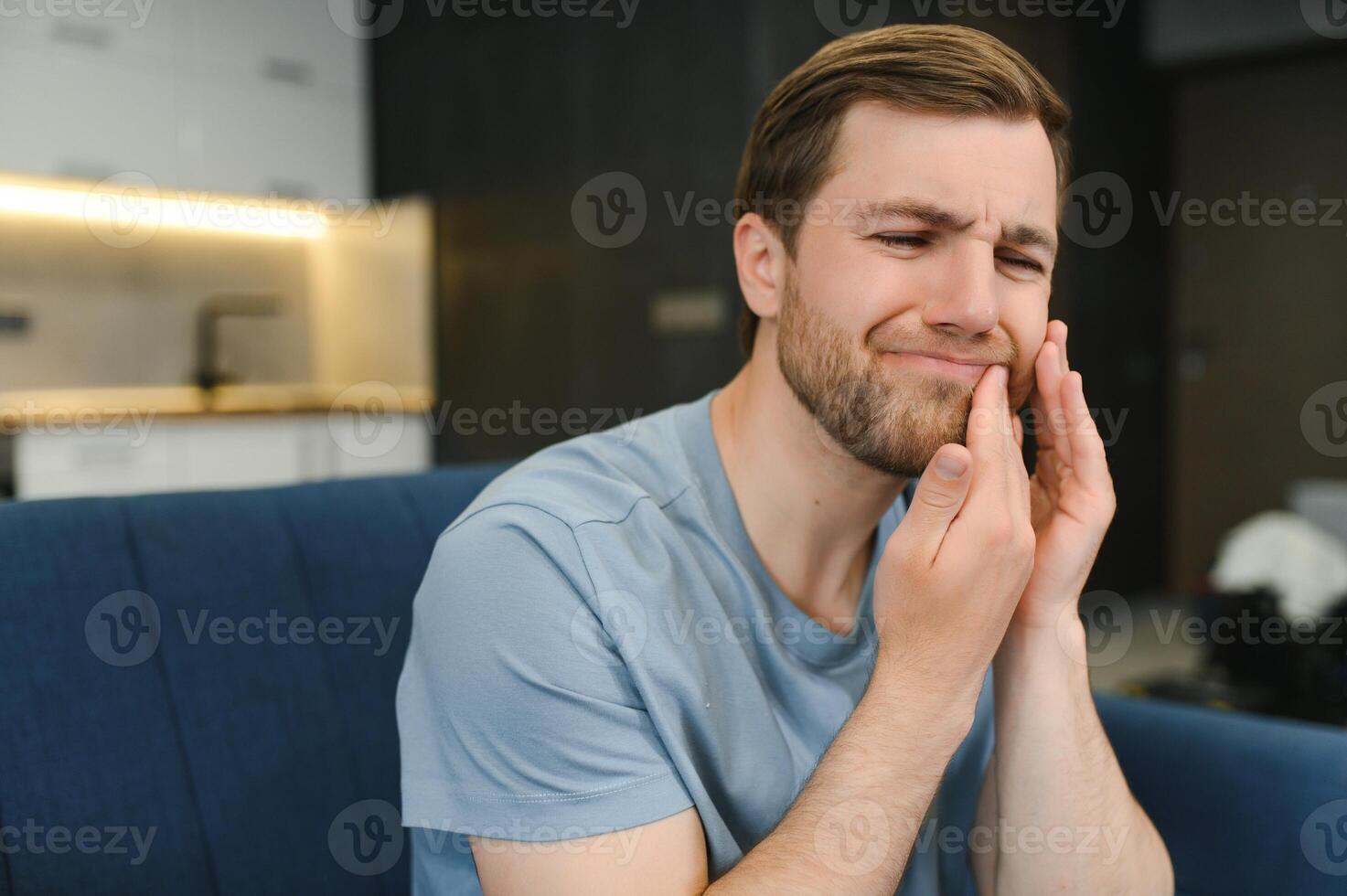 Toothache. Unhappy upset caucasian sad man sits on the couch at home, holds his hand near his cheek, grimaces, has an acute toothache, needs a consultation with a dentist doctor photo