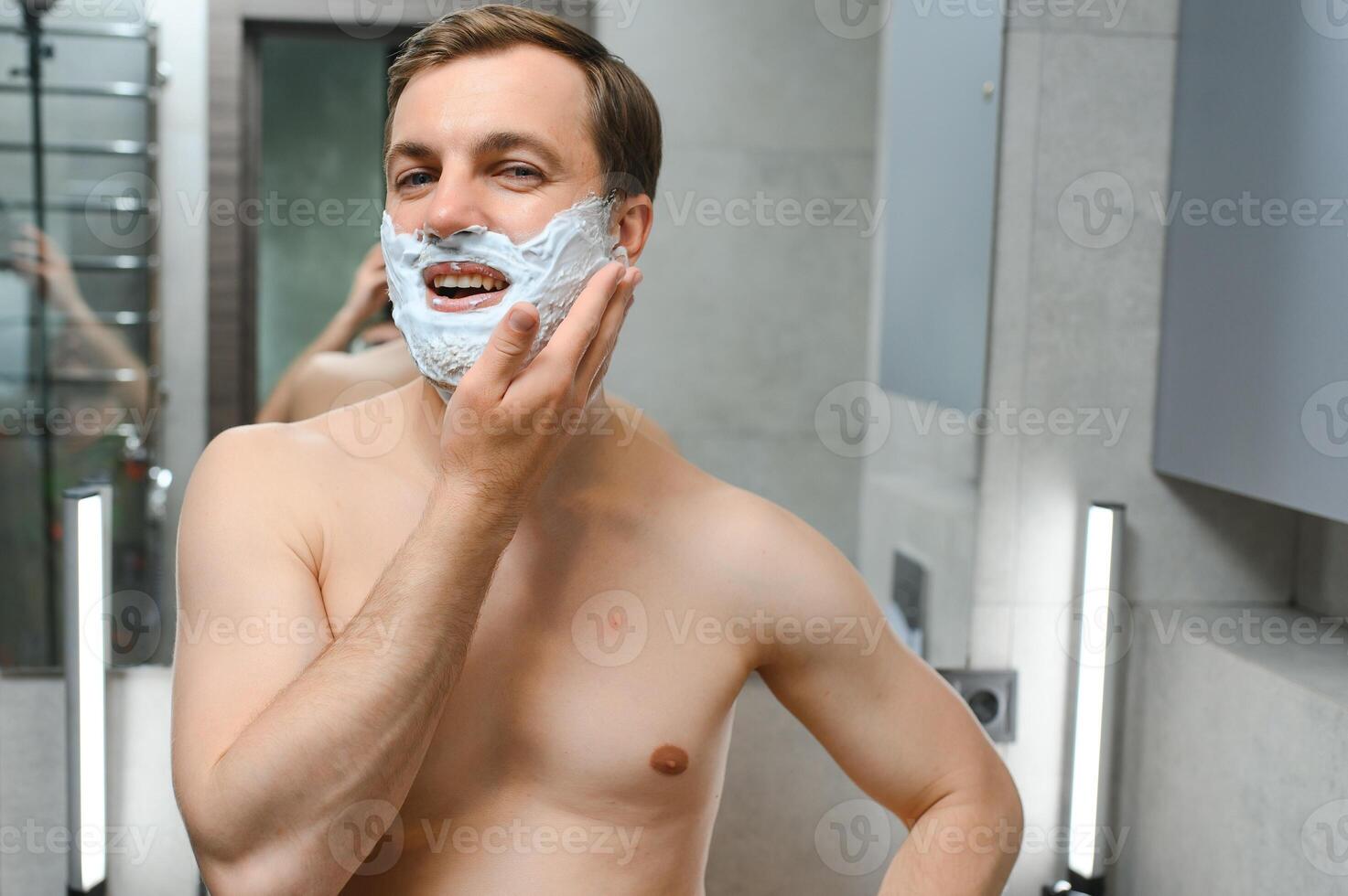 Young smiling Caucasian shirtless man applying shaving foam on face in front of mirror, preparing for hair removal morning routine. Concept of personal hygiene, skincare, male facial beauty products. photo
