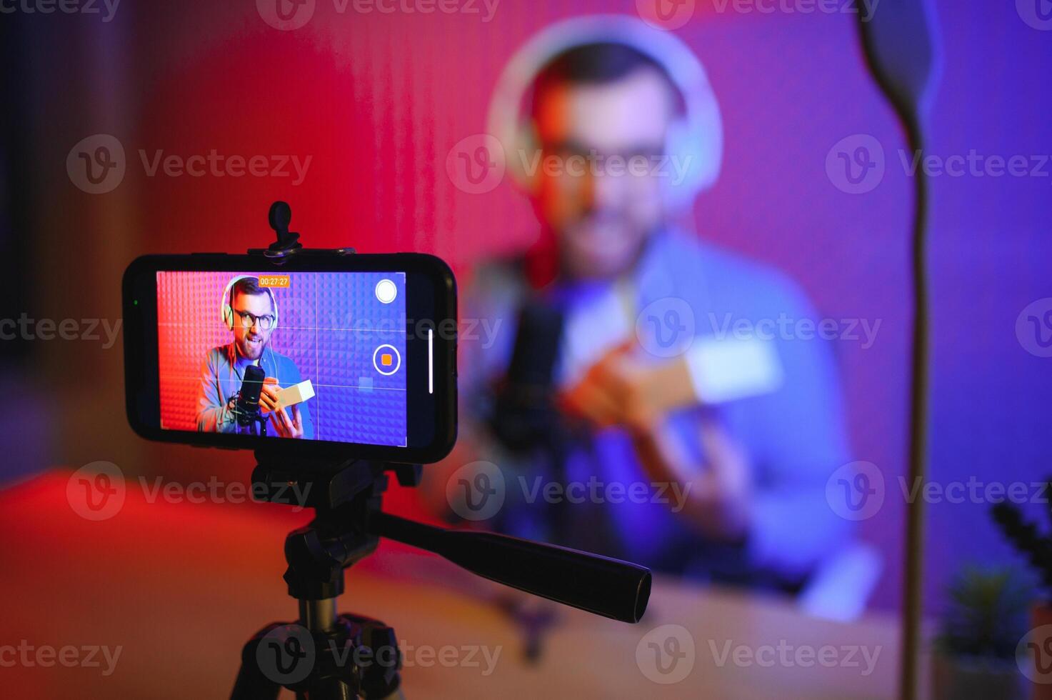 Young male technology blogger recording video blog or vlog about new smartphone and other gadgets at home studio. Blogging, Work from Home concept. Focus on gadgets at the table. Web Banner photo