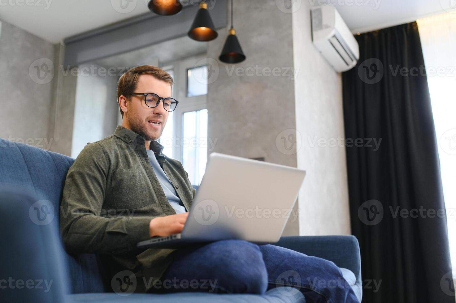 young freelancer man in stylish glasses working at laptop computer, sitting on floor at home, using wireless technology for job, leisure on Internet, thinking, touching chin photo