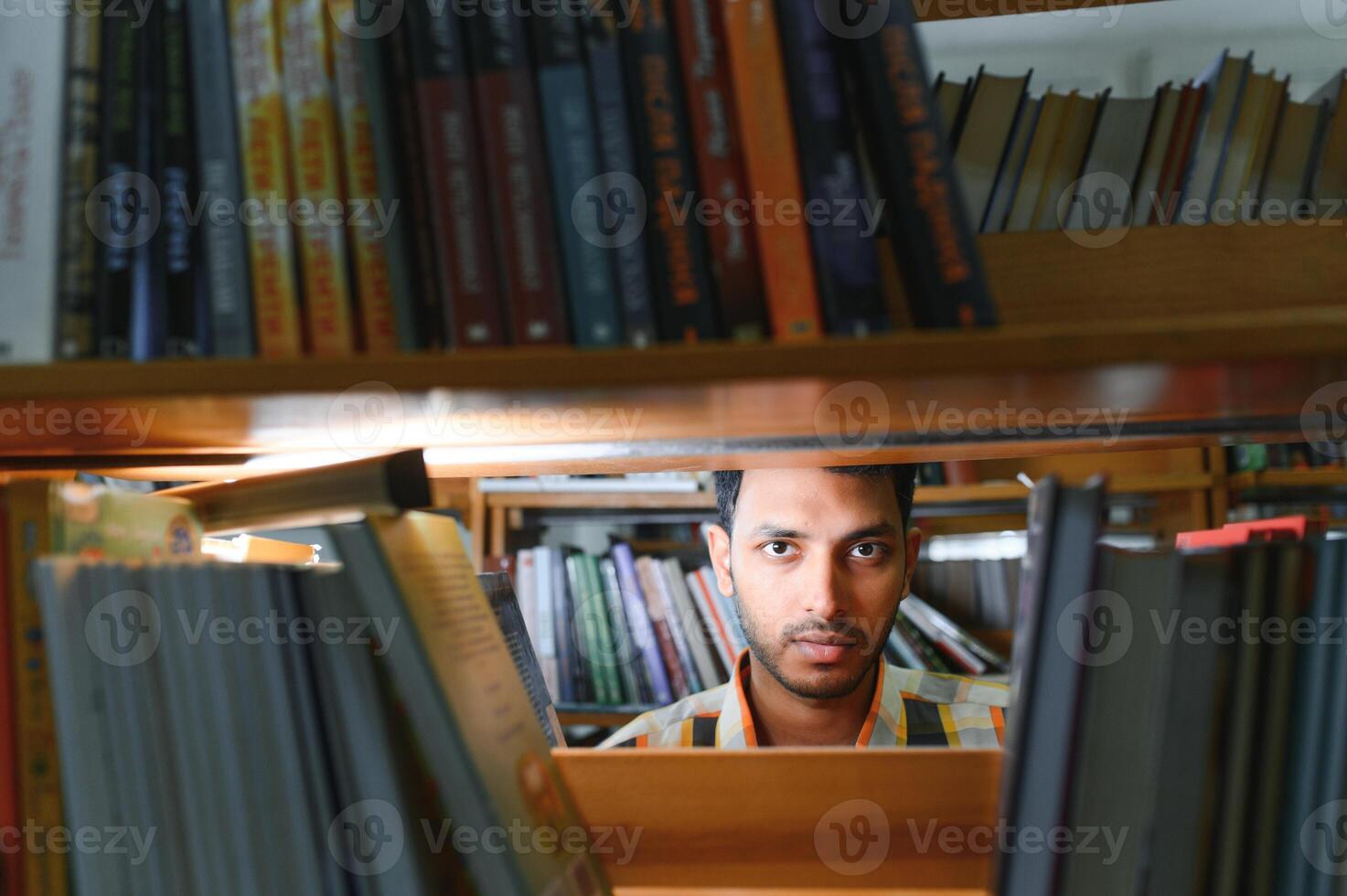 Portrait of cheerful male international Indian student with backpack, learning accessories standing near bookshelves at university library or book store during break between lessons. Education concept photo