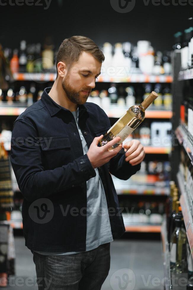 Portrait of young glad positive male customer selecting wine in supermarket photo