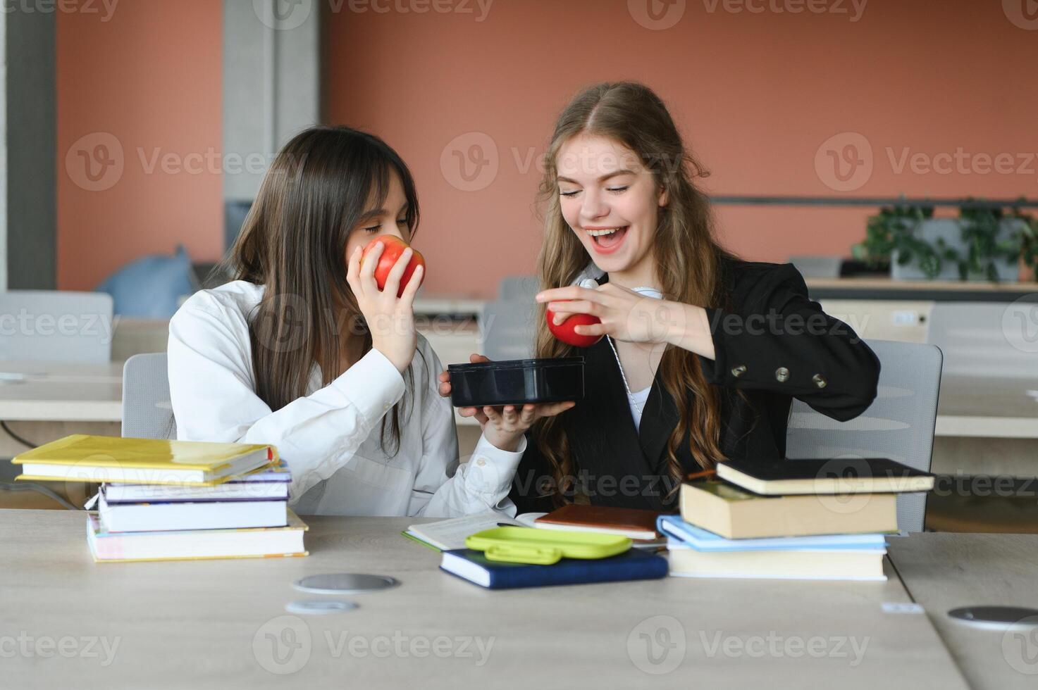 un linda niña sonriente a su compañera de clases mientras teniendo almuerzo a el escritorio en colegio durante un descanso foto